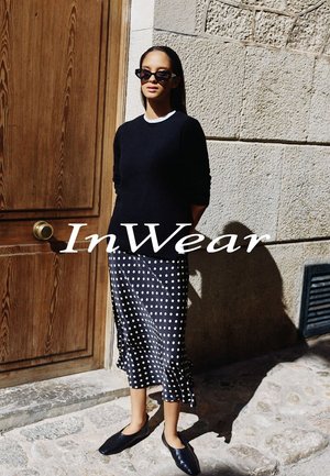 Schwarzer Pullover, Rock mit Punkten, schwarze Slipper. Glatte Texturen, figurbetonte Silhouette und Kontrast zwischen dunklem Stoff und weißen Punkten.