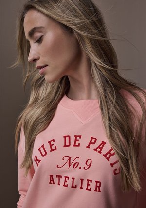 Lyserød sweatshirt med rød tekst "RUE DE PARIS No.9 ATELIER." Har rund halsudskæring og tekstureret stof, båret af en person med langt hår.