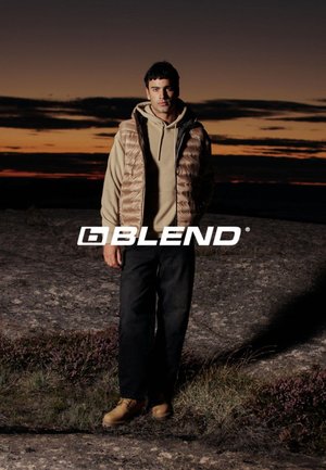Mand i beige hættejakke, tan oppustet vest, sorte bukser og tan støvler, der står på klippegrund ved solnedgang med "Blend" logoen tværs over midten.