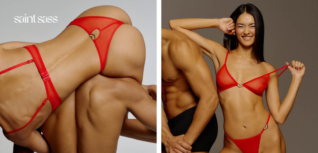 Frau in roter Unterwäsche, die spielerisch ihren Träger zieht, steht neben einem oberkörperfreien Mann in schwarzer Unterwäsche vor neutralem Hintergrund.