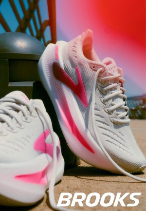 Witte Brooks hardloopschoenen met roze accenten liggen op zanderige grond bij een zwart metalen paal onder fel zonlicht.