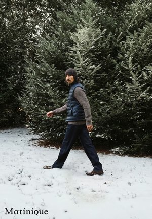Navy puffer vest over a textured gray sweater, dark pants, and brown footwear. Snowy ground and evergreen trees in the background.