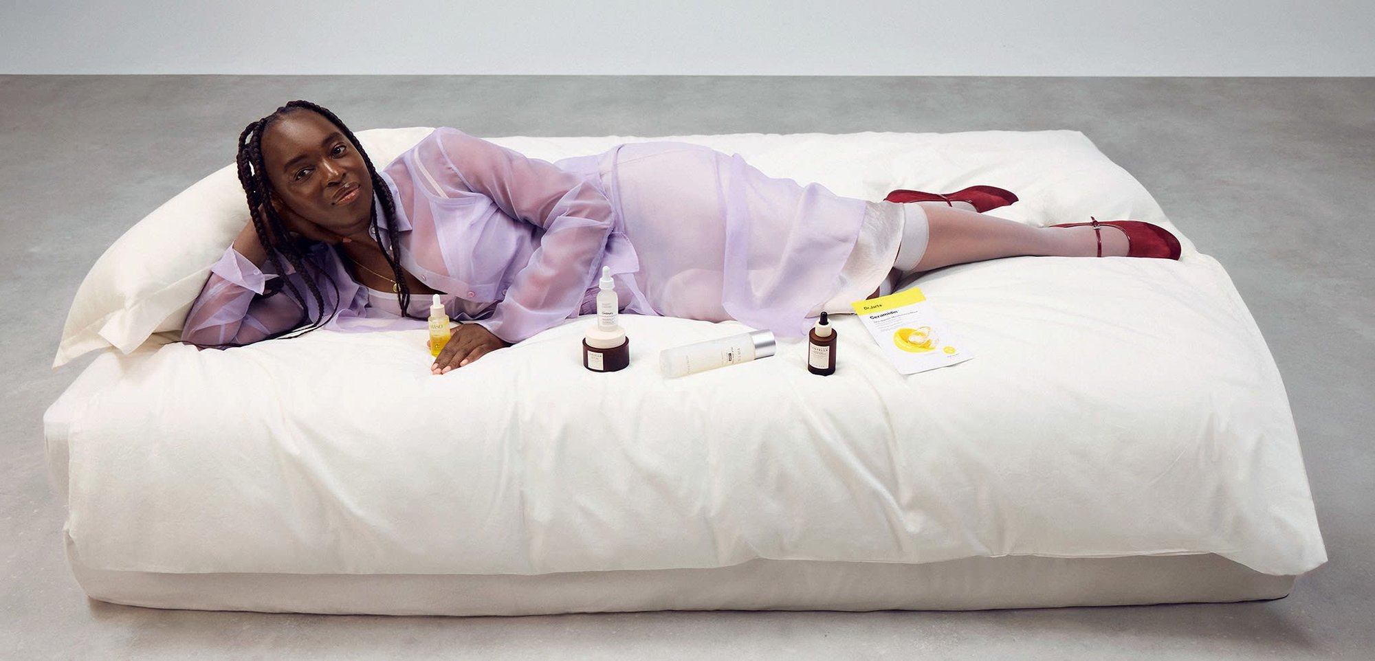 Paarse sheer ochtendjas, witte aansluitende kleding eronder, liggend op een groot wit kussen. Huidverzorgingsproducten en een gele verpakking zijn in de buurt. Rode schoenen.