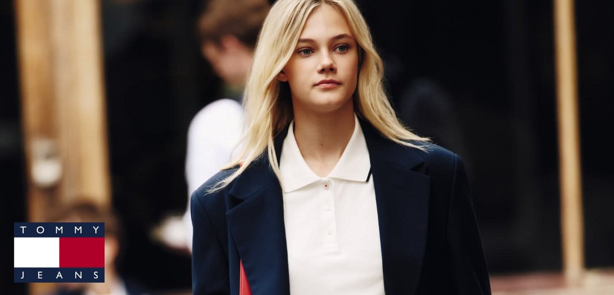 Jeune femme aux longs cheveux blonds portant une chemise blanche à col et un blazer sombre, marchant en plein air, avec le logo Tommy Jeans dans le coin.