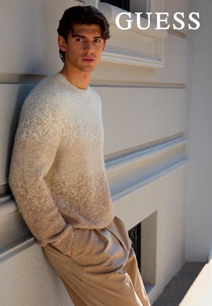 Maglione sfumato beige e crema, con una texture morbida e soffice. Il modello lo indossa abbinato a pantaloni beige chiaro, in piedi contro una parete neutra.
