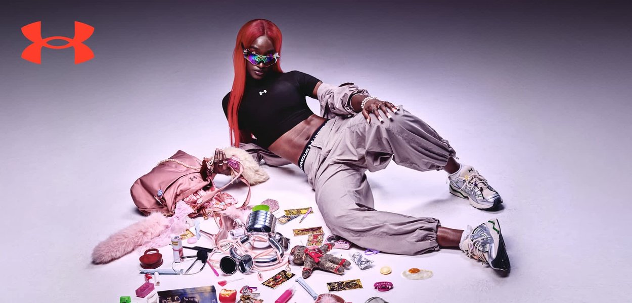 Person mit langen pinkfarbenen Haaren und Kapuzenweste hockt, umgeben von Boxhandschuhen, Buch, Sportbekleidung und verstreuten kleinen Gegenständen auf weißem Boden.