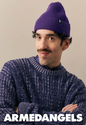 Paarse geribde beanie en een gestructureerde marineblauwe trui met een brede halslijn en een ingewikkeld patroon. Model poseert met gekruiste handen.
