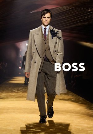 Męski model maszerujący po wybiegu w beżowym płaszczu, brązowym garniturze, paskowanej koszuli, bordo krawacie i czarnych butach z napisem "BOSS" po prawej stronie.
