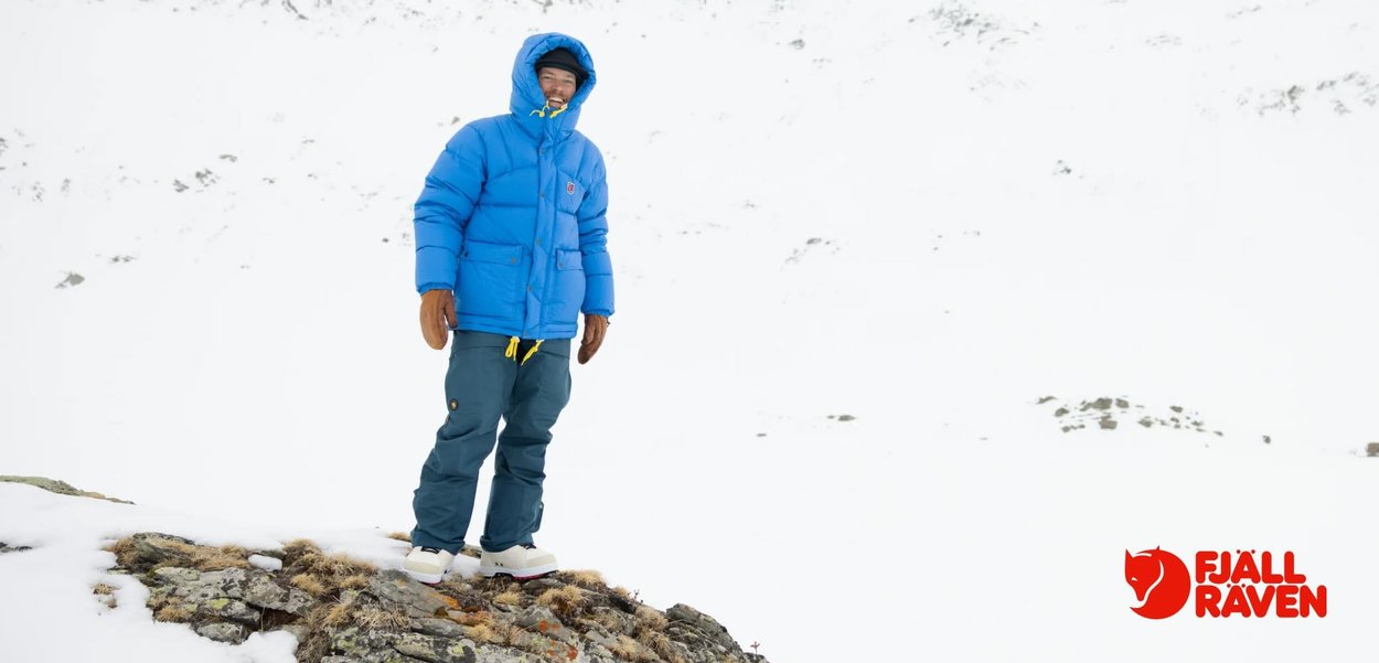 Blå vadderad jacka, gröna byxor, bruna handskar och vita stövlar. Står på en stenig yta med snö och en vit bergig bakgrund.