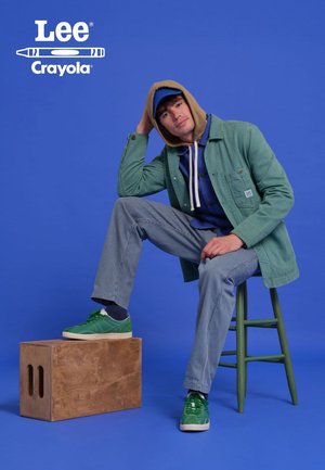 Green corduroy jacket layered over a blue hoodie, striped pants, and green sneakers. Wooden box beside, bright blue background.