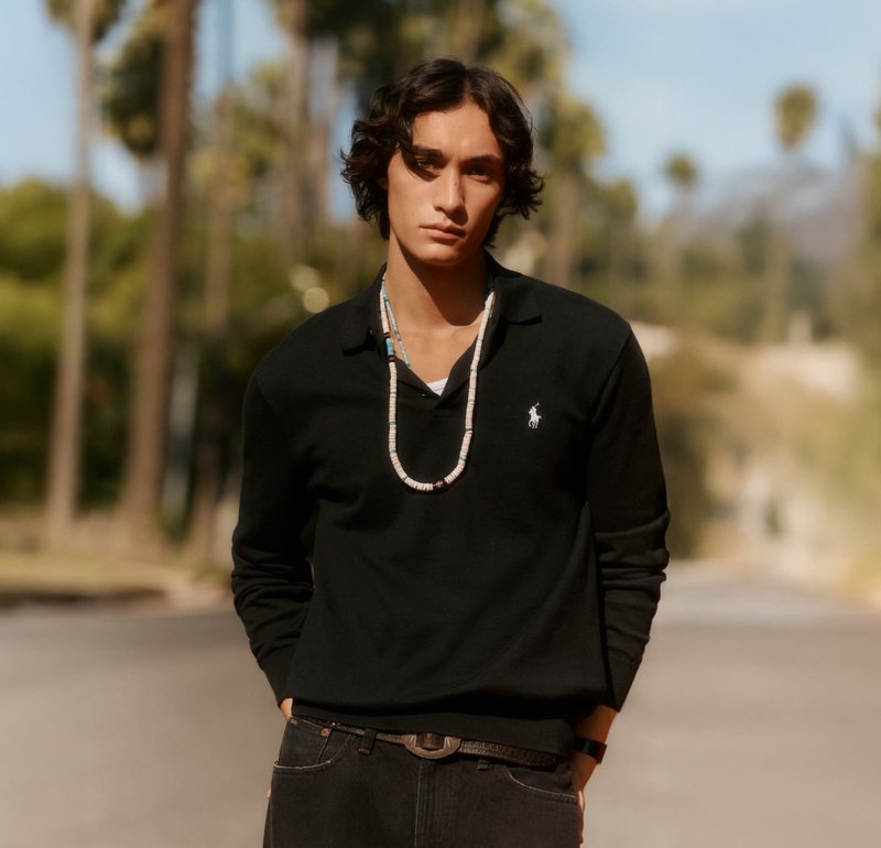 Joven con cabello de longitud media que lleva una camiseta negra de manga larga y un collar de cuentas, de pie al aire libre con las manos en los bolsillos.