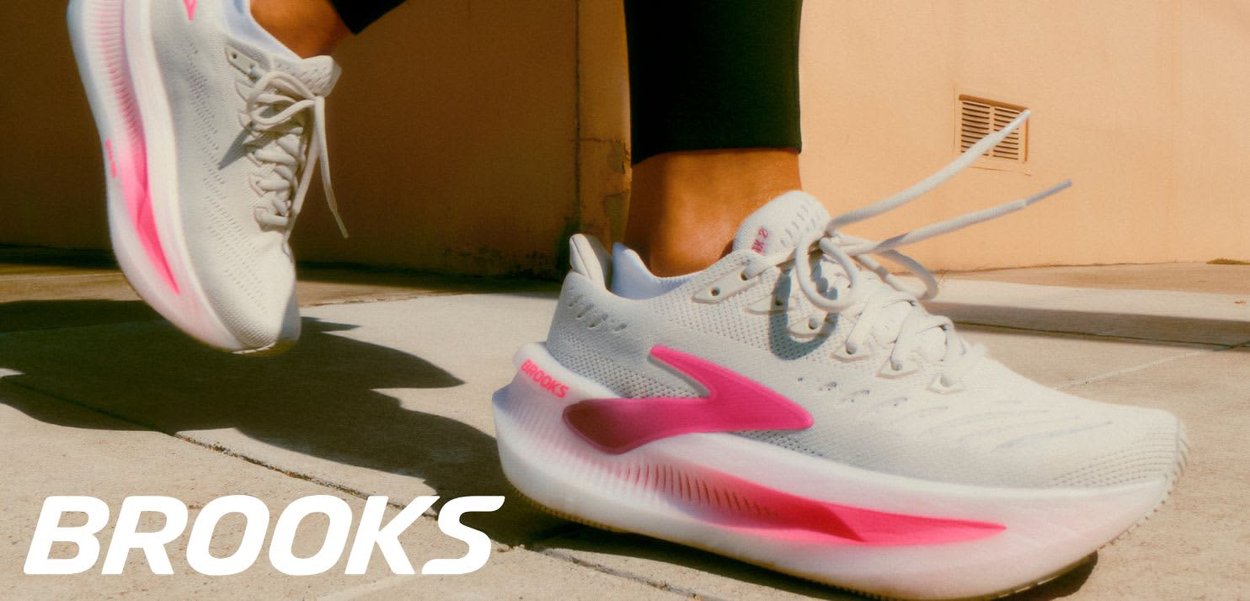 Persoon met witte en roze Brooks hardloopschoenen loopt op zonovergoten trottoir bij een beige muur, met een voet halverwege in de lucht.