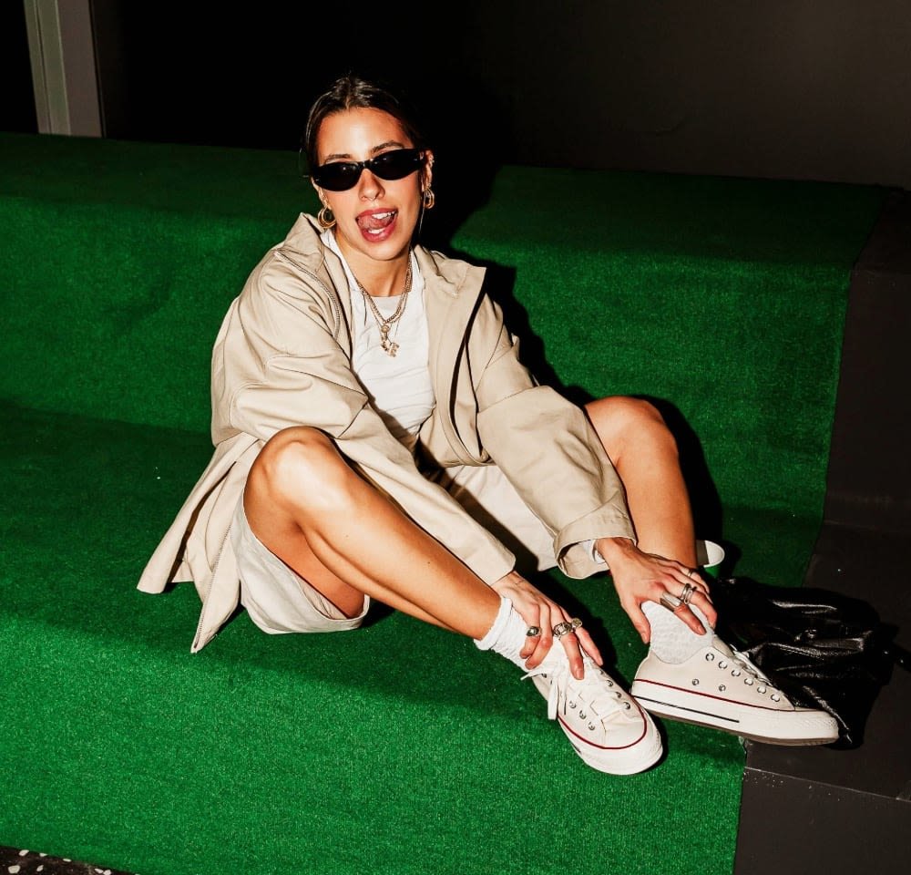 Beige trench coat over white top, seated on green steps. Wearing black sunglasses and gray/white sneakers, accessorized with rings.