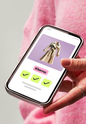 Beige Trenchcoat auf einem Smartphone vor lila Hintergrund präsentiert. Die Benutzeroberfläche zeigt Klarna-Branding und Häkchen-Symbole.