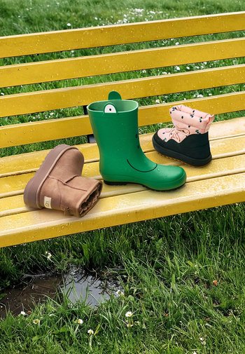 Kolm paari saapaid kollasel pingil: rohelised kummist vihmasaapad, beežid faux karvajopega ankle saapad ja mustad-roosad veekindlad saapad lillemustriga.