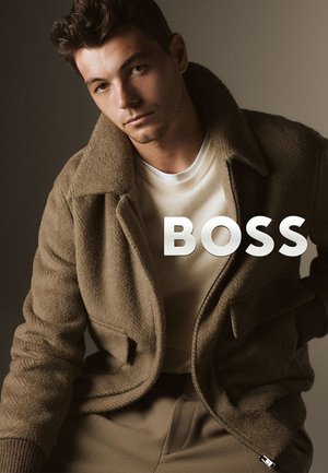 Giacca in pile marrone con ampio colletto, sopra una camicia color crema. Texture morbida, chiusura con bottoni e tasche laterali visibili. Logo "BOSS" in bianco.