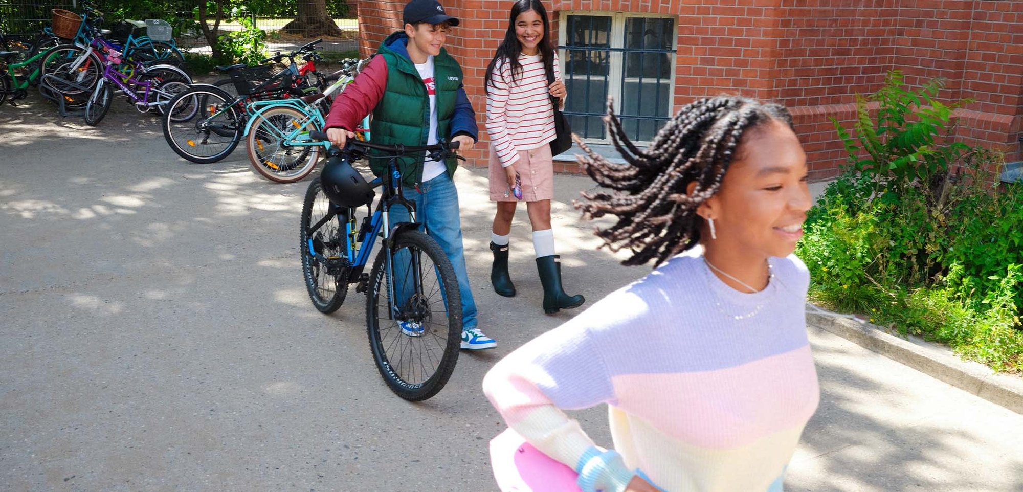 Trois enfants souriants à l'extérieur : un pousse un vélo, un marche avec des bottes de pluie, et un court avec des cheveux tressés flottants.