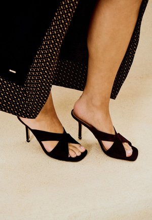Pies con mules de tacón alto de ante negro con tiras cruzadas, combinados con una falda larga estampada en un suelo claro.