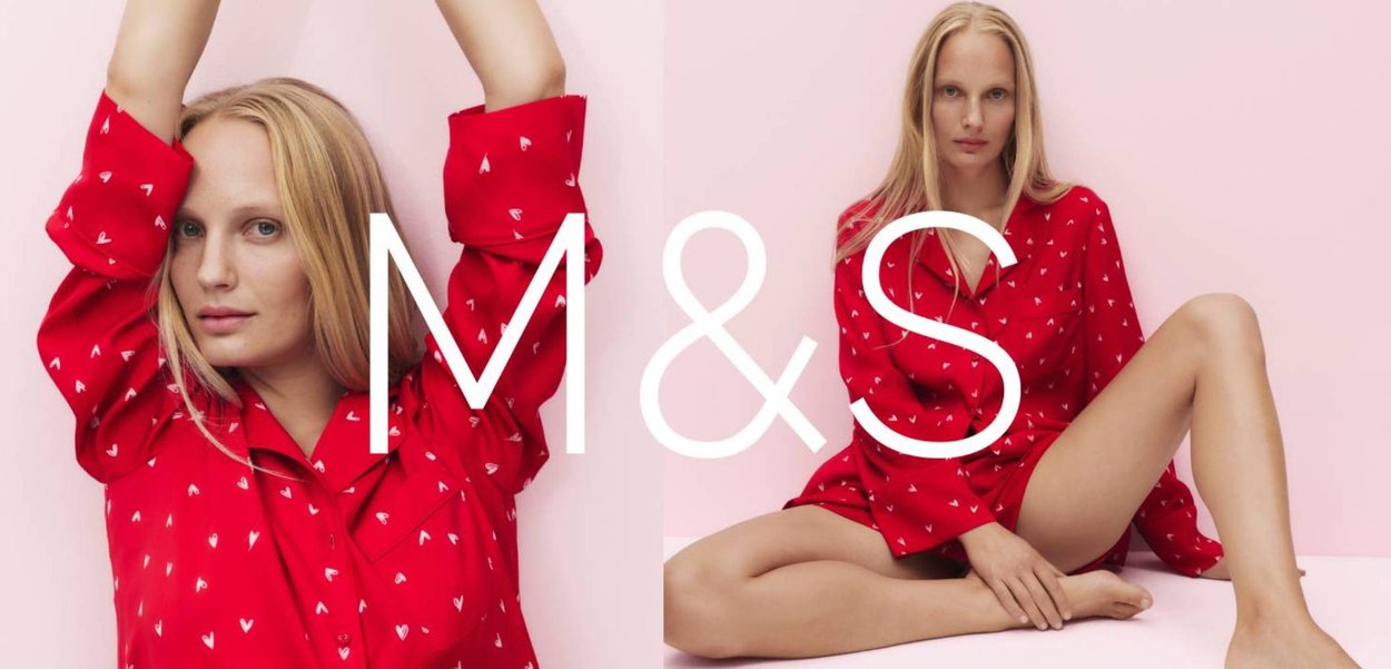 Femme blonde portant une chemise rouge à motifs de cœurs blancs, posant les bras levés devant un fond rose pâle.