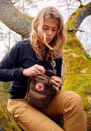 Brun Fjällräven Kanken mini-väska med röd logotyp, med en framficka och justerbar rem, hålls av en person i en svart tröja.