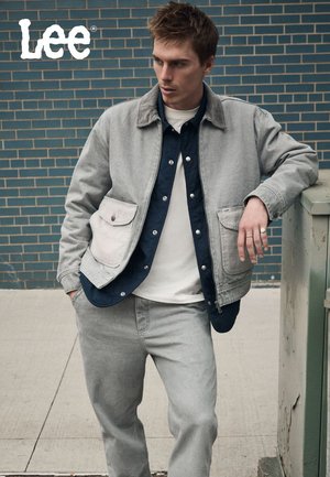 Chaqueta de mezclilla gris sobre una camisa oscura con botones, combinada con pantalones gris claro. La chaqueta tiene bolsillos delanteros y textura contrastante.