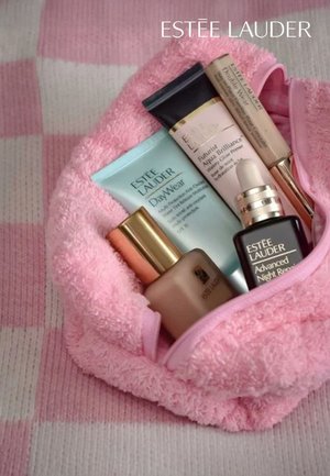 Pink terry cloth bag contains five Estée Lauder skincare products in various shapes; notable items include a black dropper bottle and a light blue tube.