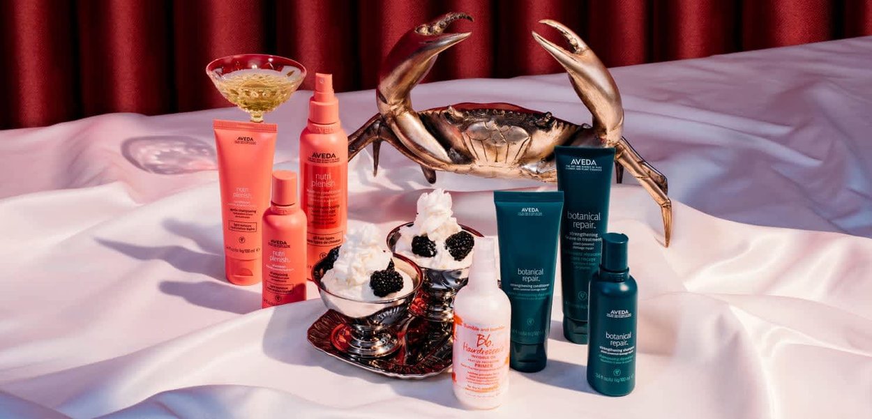 Prodotti per la cura dei capelli Aveda in confezioni vivaci rosa e verde acqua, disposti insieme a una scultura di granchio e ciotole di crema guarnite con more.