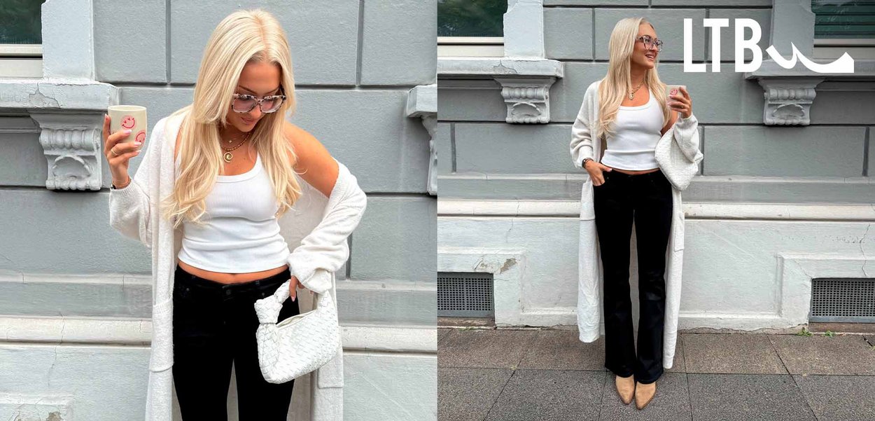 Witte lange cardigan over een aansluitend wit tanktop, gecombineerd met zwarte flare jeans en beige puntige hakken. Houdt een roze beker vast.