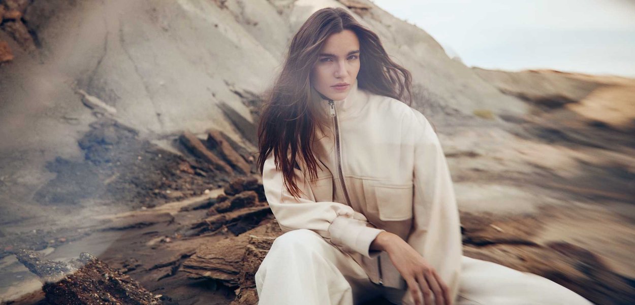 Młoda kobieta o długich brązowych włosach ubrana w beżową kurtkę i spodnie, siedzi na skalistym terenie, o miękkim naturalnym oświetleniu i neutralnych tonach.
