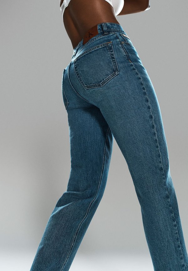 Jeans de mezclilla azul con un corte ajustado, costuras visibles, cinco bolsillos y un parche de cuero en la cintura. La textura parece suave y duradera.