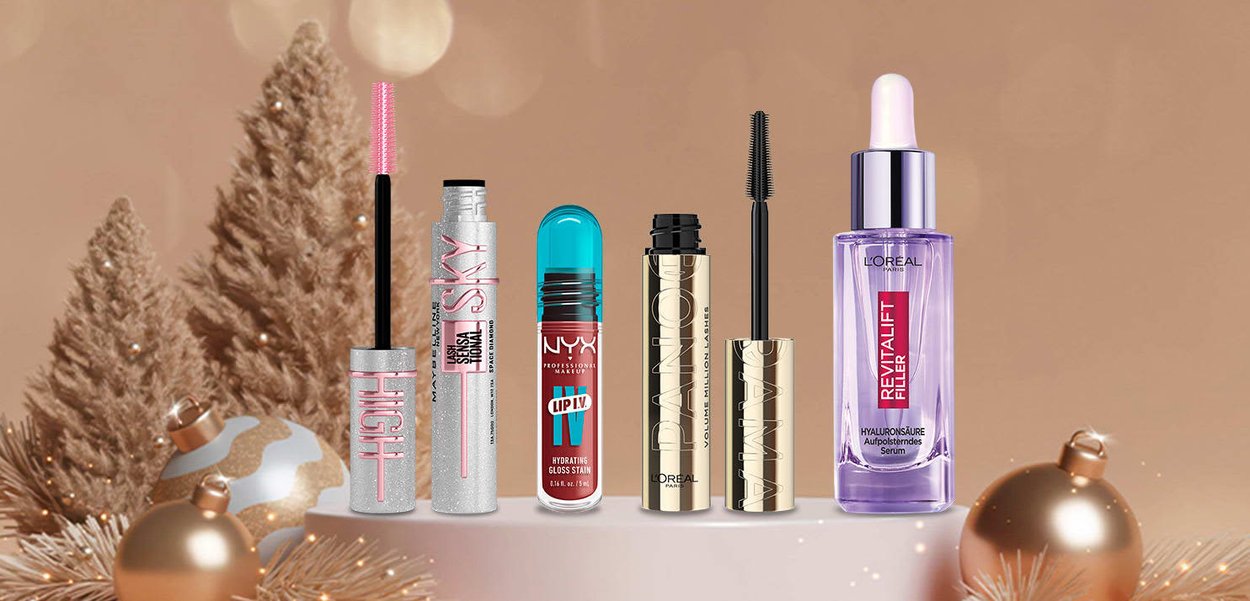 Sei prodotti cosmetici disposti in fila: un tubo di mascara glitterato, una tinta labbra turchese, un mascara dorato e una bottiglia di siero viola.