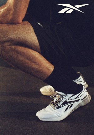 Persoon in zwarte korte broek en sokken die een squat doet, draagt witte en zwarte Reebok sportschoenen met zichtbaar logo.