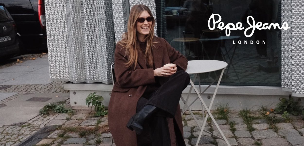 Abrigo marrón y pantalones negros llevados por una persona sentada en una pequeña mesa redonda. El fondo presenta una cortina texturizada de color gris claro y adoquines.