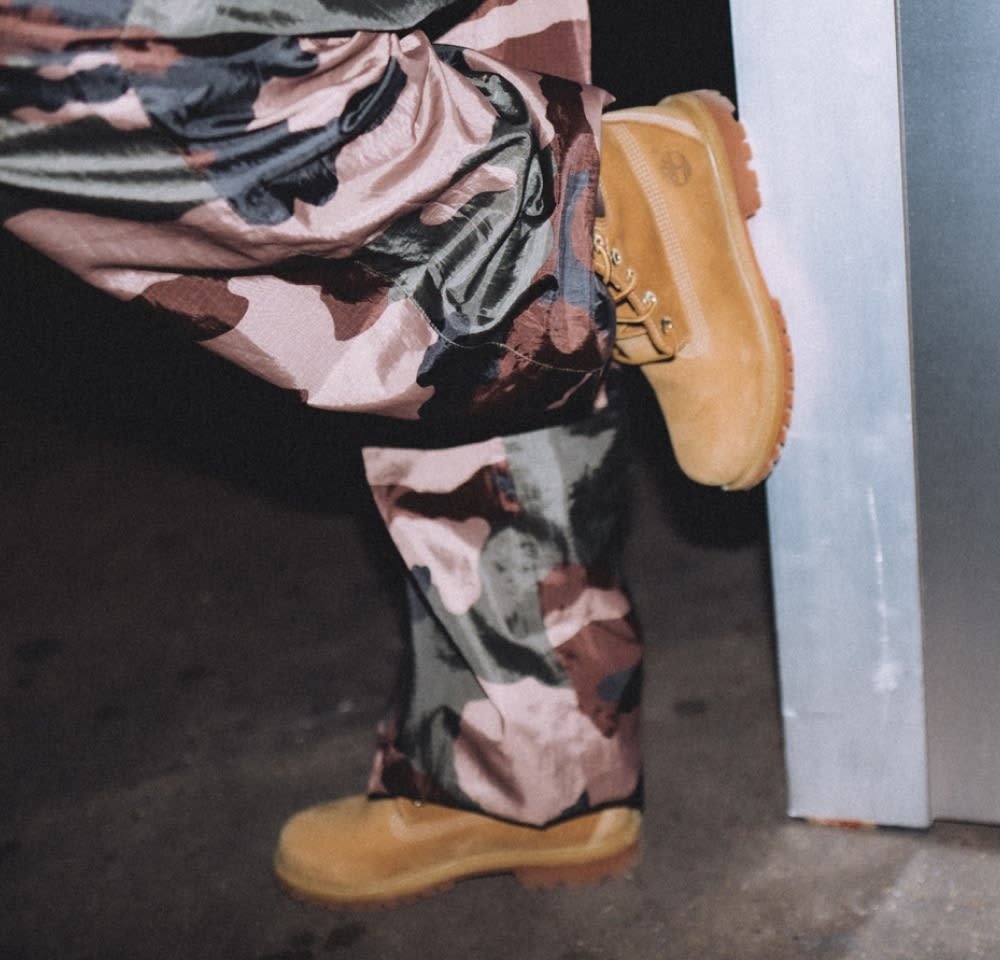 Tan wasserfeste Stiefel mit strukturierten Sohlen und goldenen Schnürsenkeln, kombiniert mit bunten, camo-musternden Hosen in den Farbtönen Rosa, Grün und Blau.