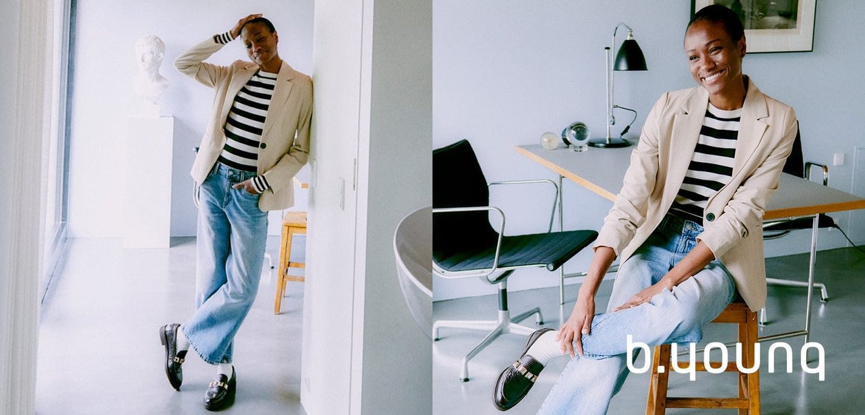 Blazer beige sur un haut rayé noir et blanc, associé à un jean bleu clair et des mocassins brillants, dans un environnement de bureau moderne.