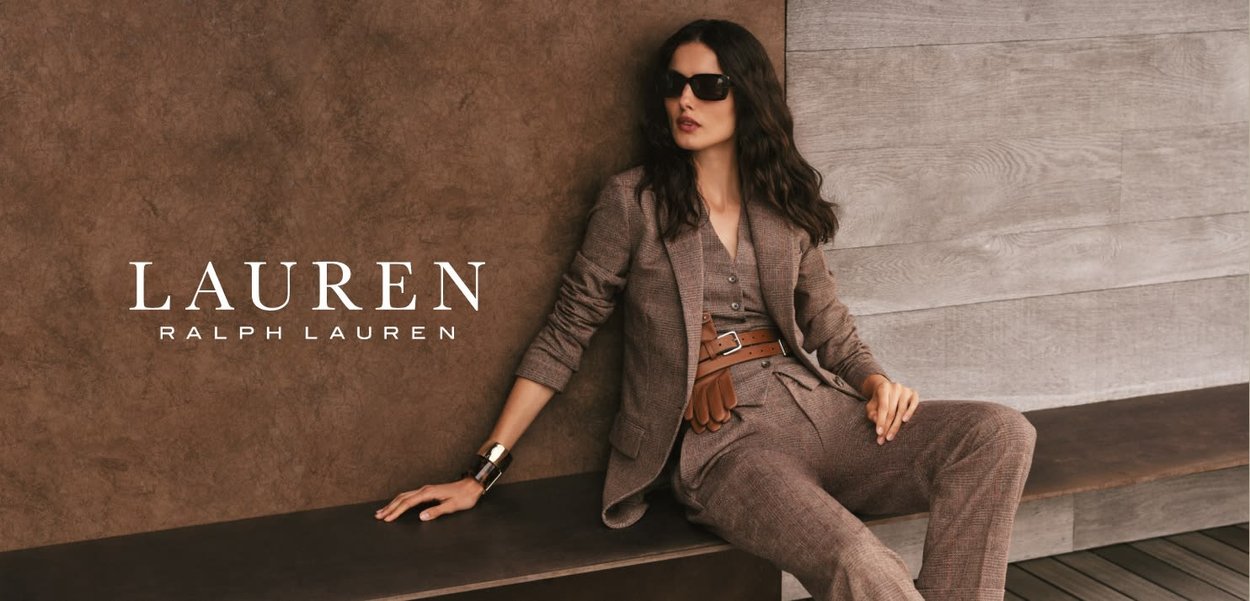 Bruin geruit pak met een getailleerde jas en bijpassende broek. Model draagt een zonnebril en een leren riem, gezeten tegen een gestructureerde achtergrond.