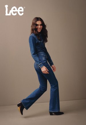 Denim jumpsuit med lange ermer, utsvingte ben og lommer foran; mørk blå farge; stylet med svarte ankelstøvletter og bølget hår.