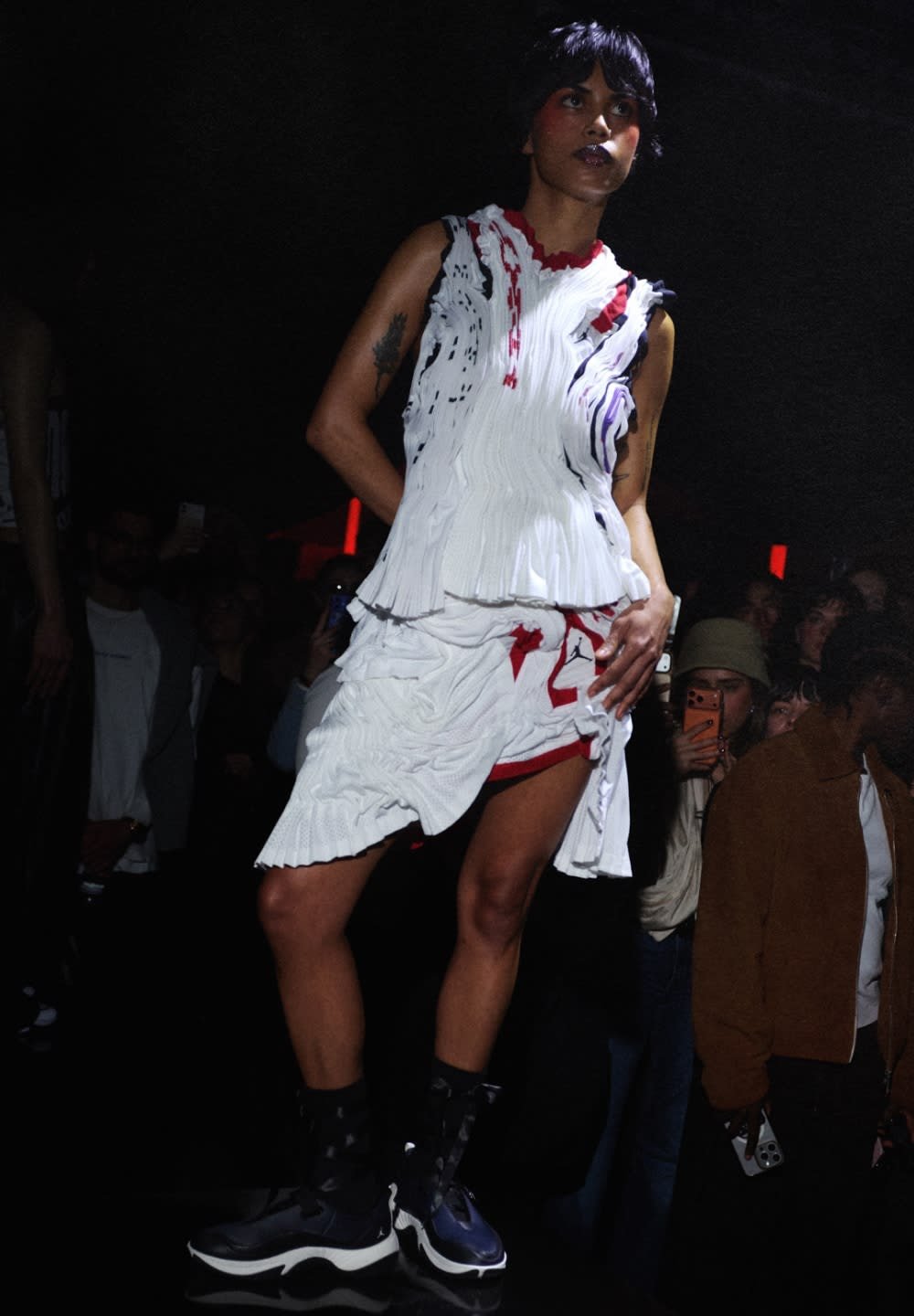 Modelo vistiendo un vestido blanco texturizado con detalles en rojo y negro, combinado con zapatos deportivos negros, en una pasarela con poca luz y audiencia al fondo.