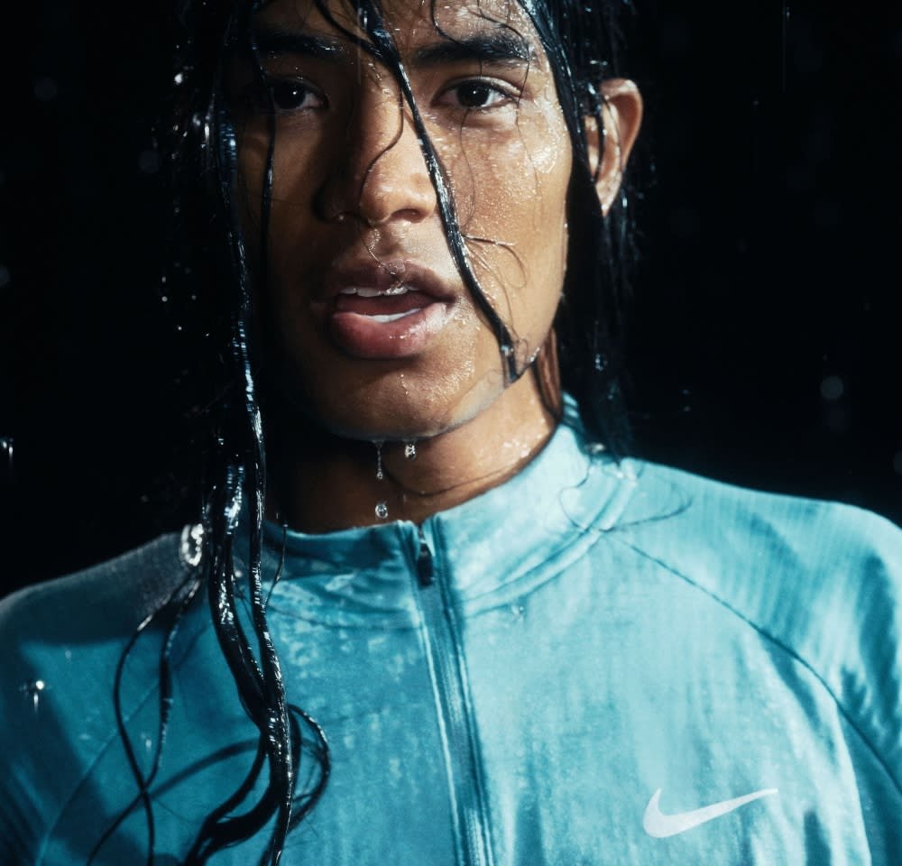 Chemise de sport zippée bleue avec une finition mate et un logo Nike blanc. Gouttes d'eau sur la peau et les cheveux, fond sombre.
