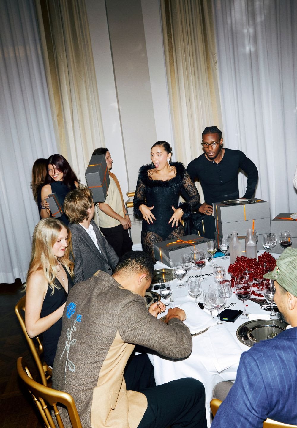 Gruppe af mennesker ved et spisebord med bestik og vinglas. Nogle holder grå kasser med orange accenter; en kvinde i en sort kjole med fjer.