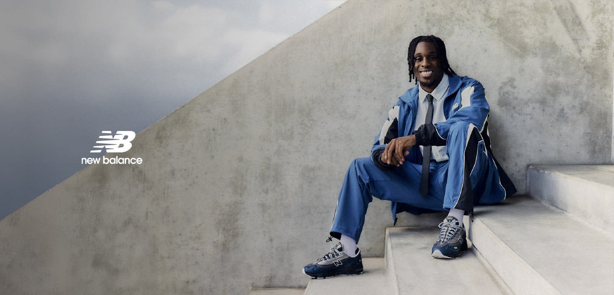 Uomo sorridente in tuta blu e cravatta seduto su gradini di cemento, con sneakers New Balance, contro un muro di cemento grigio.
