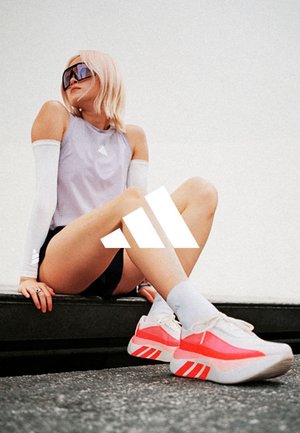 Joven mujer con gafas de sol, ropa deportiva y zapatillas Adidas de color rosa brillante y blanco sentada en un borde bajo al aire libre.