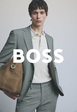 Light gray blazer and trousers with a textured off-white polo shirt. Brown leather bag with silver hardware is held in the left hand.