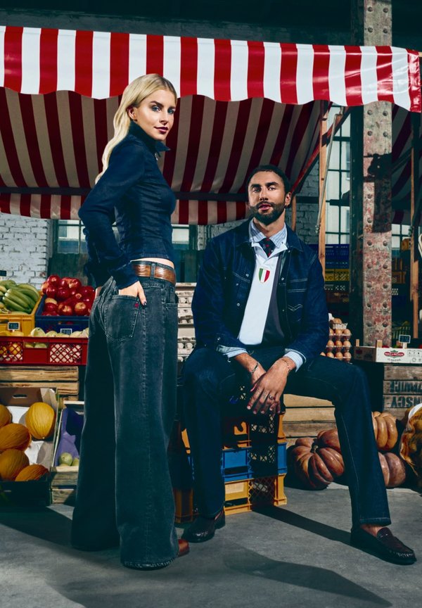 Giacca di jeans e camicia a maniche lunghe su modello; jeans grigi a gamba larga. Uomo in giacca scura con camicia blu, colletto a fantasia. Sfondo di un mercato.