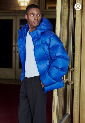 Junger Mann in leuchtend blauem Steppjacke, weißem Hemd und schwarzer Hose lehnt an einem kunstvoll verzierten goldenen Türrahmen.