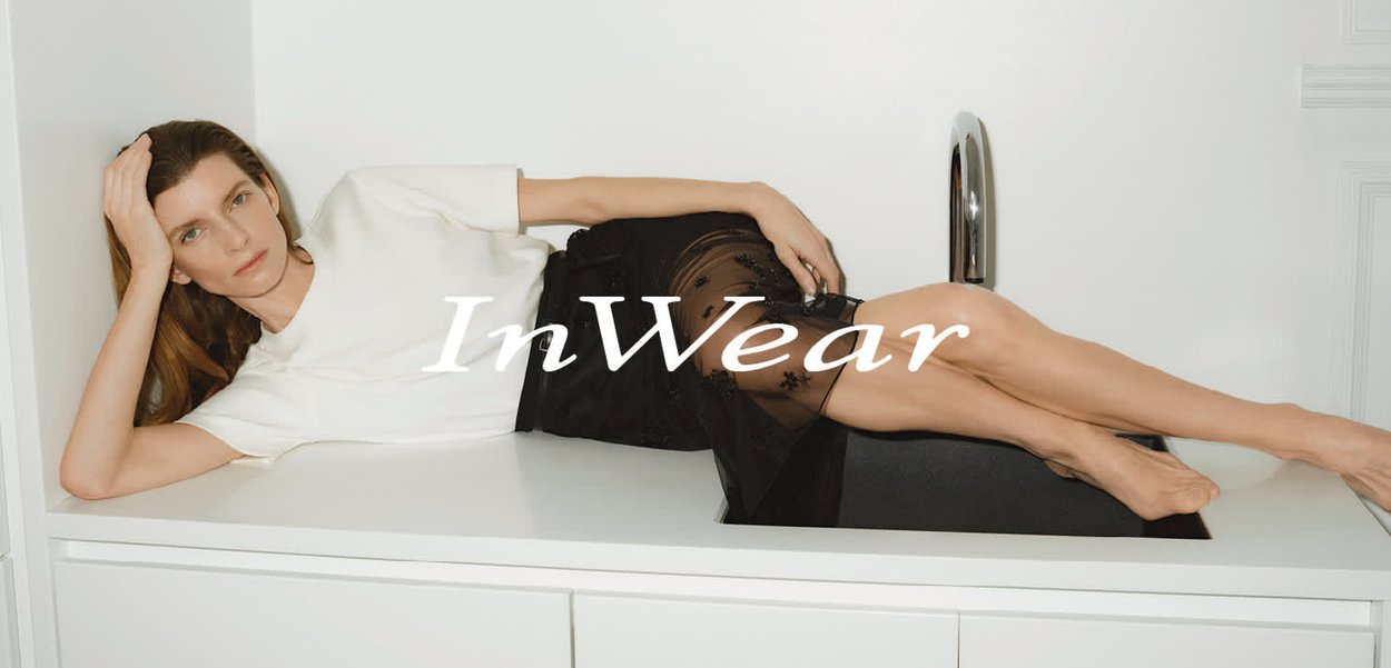 White short-sleeve top paired with a sheer black skirt featuring floral embroidery. Model reclining on a white surface with a sleek silver faucet.