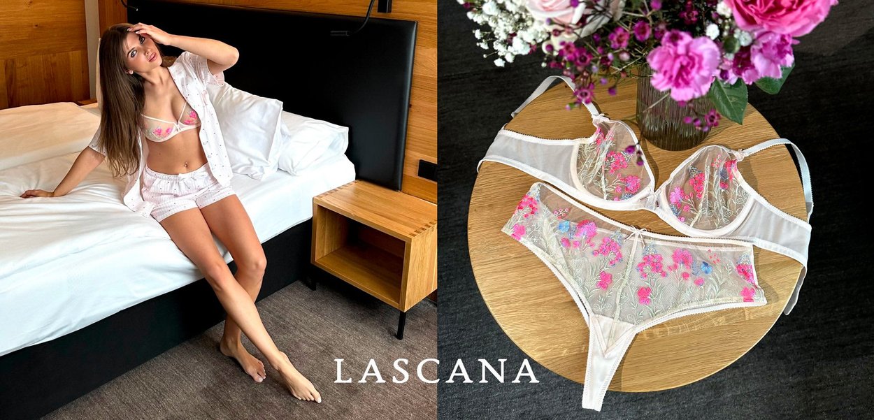 Frau mit langem braunen Haar, die beige Lingerie mit rosa Blütenstickerei trägt und an einer hellgrauen Wand neben einem durchsichtigen Vorhang steht.