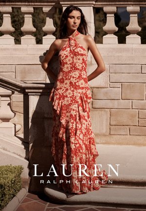 Mujer de pie junto a una balaustrada de piedra, lleva un vestido maxi con estampado floral en rojo y beige, con cuello halter y capas con volantes.