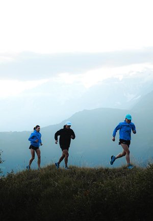 Trois coureurs sur une colline herbeuse ; l'un en veste bleue, un autre en noir, et un troisième en chemise et casquette bleues. Des montagnes en arrière-plan.