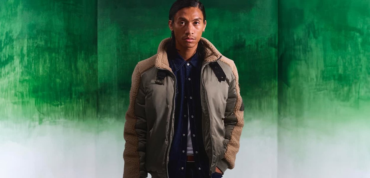 Puchowa oliwkowozielona kurtka bomberka z beżowymi akcentami z owczej wełny, z czarnym kołnierzem, zamkiem błyskawicznym z przodu i guzikami, noszona na ciemnoniebieskiej koszuli.